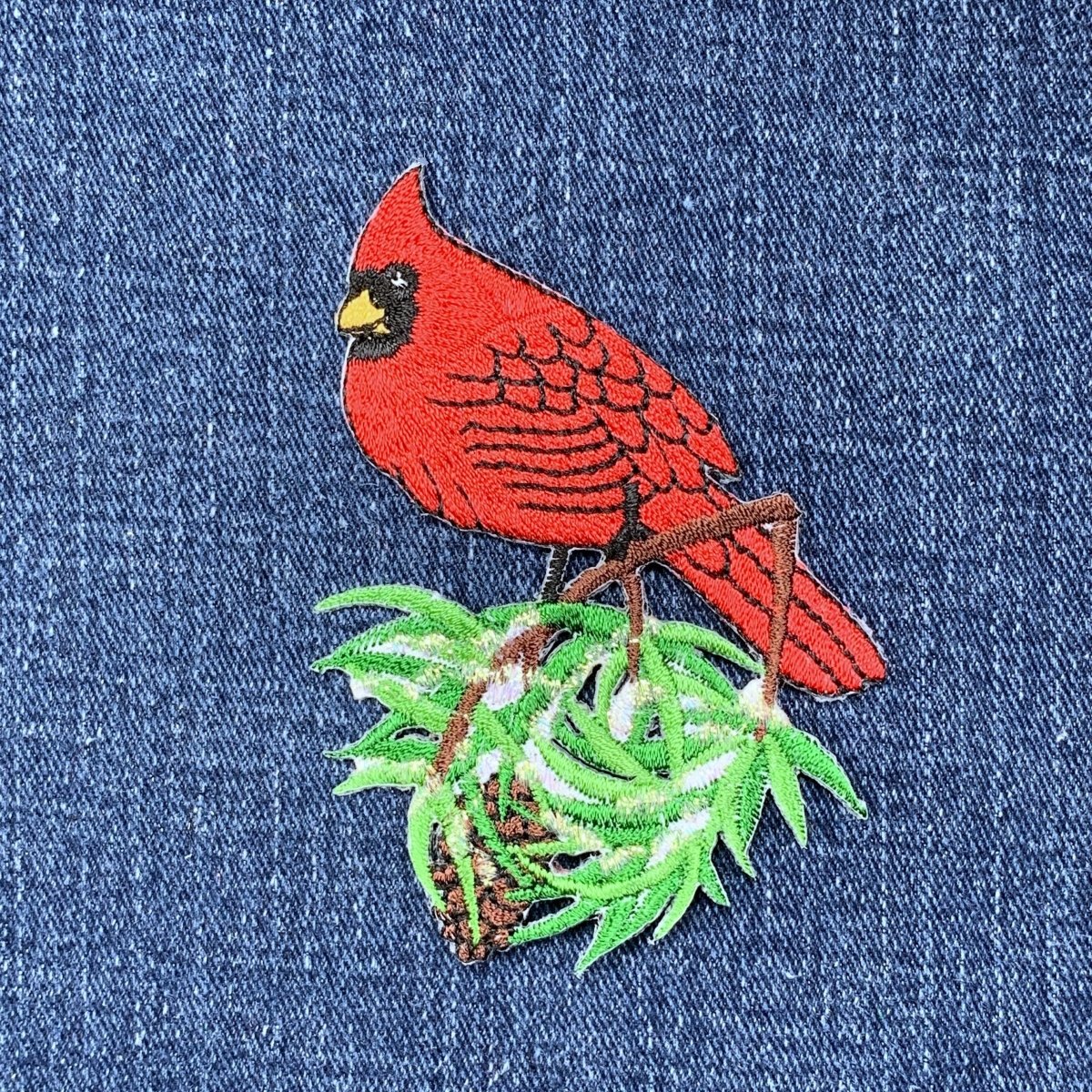 Embroidered red cardinal bird on a branch with green leaves on denim fabric.