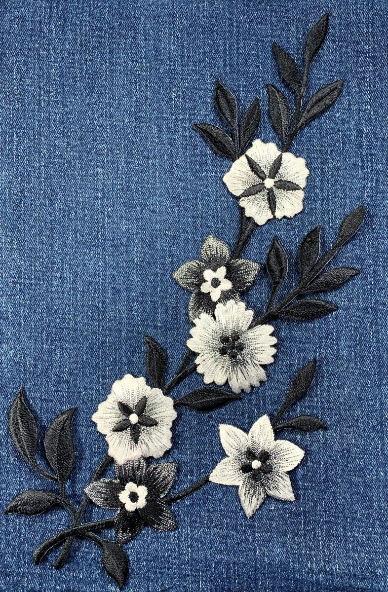 Embroidered appliqué featuring a winding vine of six flowers and several leaves, stitched in white, black, and grayscale thread, against a background of bright blue denim fabric.