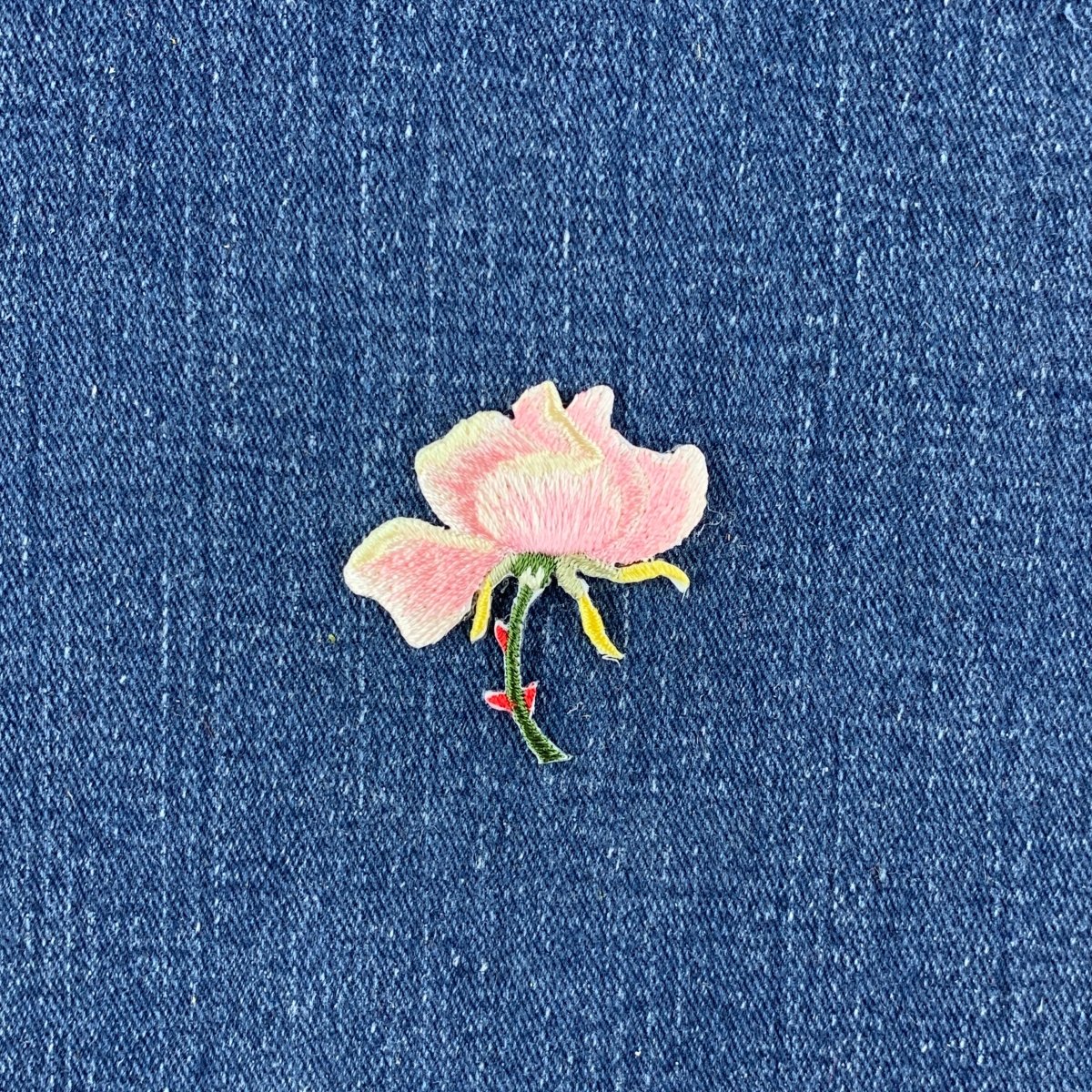 A small embroidered pink rose flower patch with yellow and green details, placed on a textured blue fabric background.