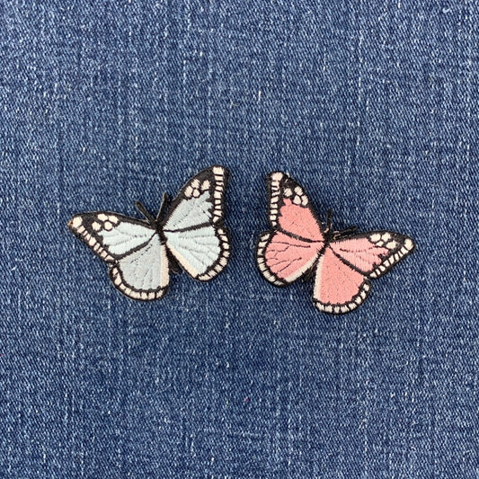 Two butterfly-shaped patches, one pale blue and one pink, with black outlines and white-tipped wings, placed on a blue denim fabric background.